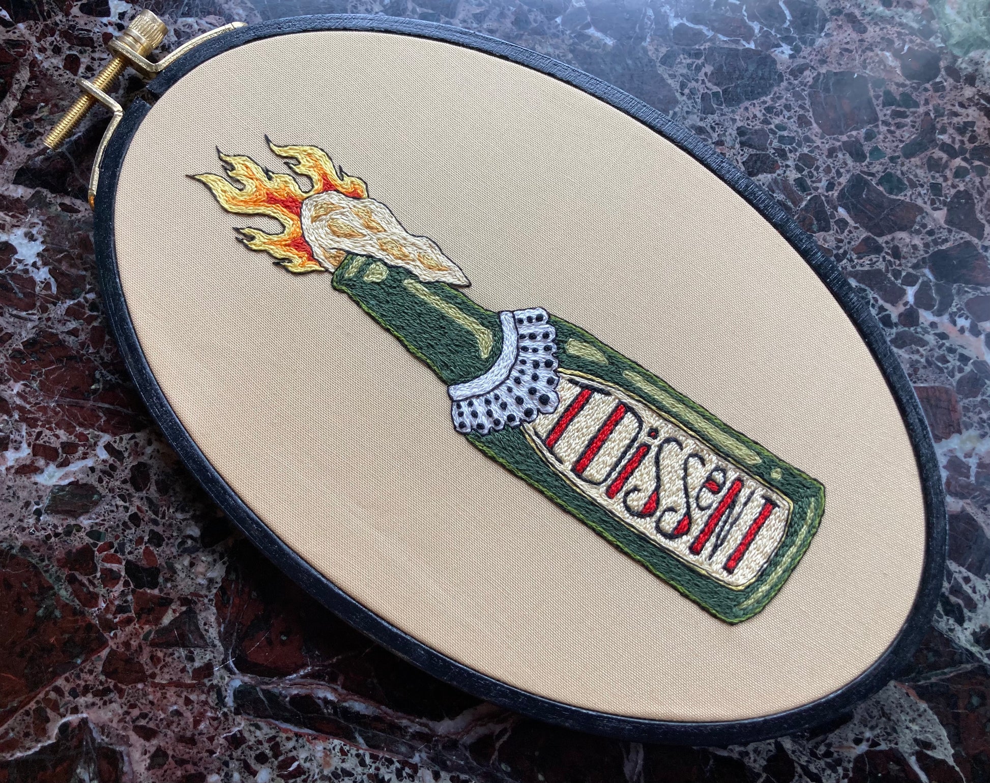 A flaming molotov cocktail bottle with a lace collar around the neck in the style of Ruth Bader Ginsburg is embroidered onto tan fabric in a black oval hoop. The label of the bottle reads "I DISSENT" in decorative font. The hoop sits upon a dark marble surface.