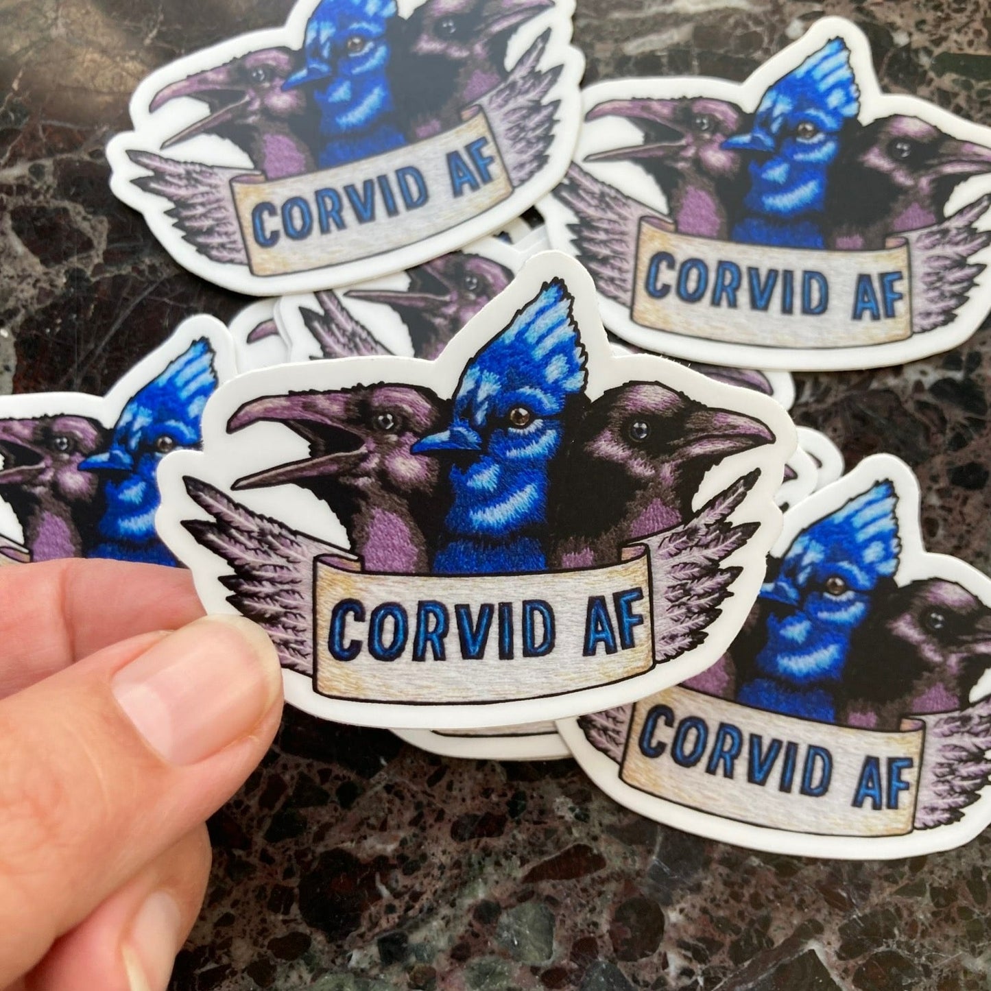 A pile of stickers sits atop a dark marble surface. The stickers depict an embroidery of the heads of three Corvids: a raven, a blue jay, and a crow. Below them is a banner that reads "CORVID AF" and the edge of the banner is feathers. a hand holds one of the stickers up to the camera.