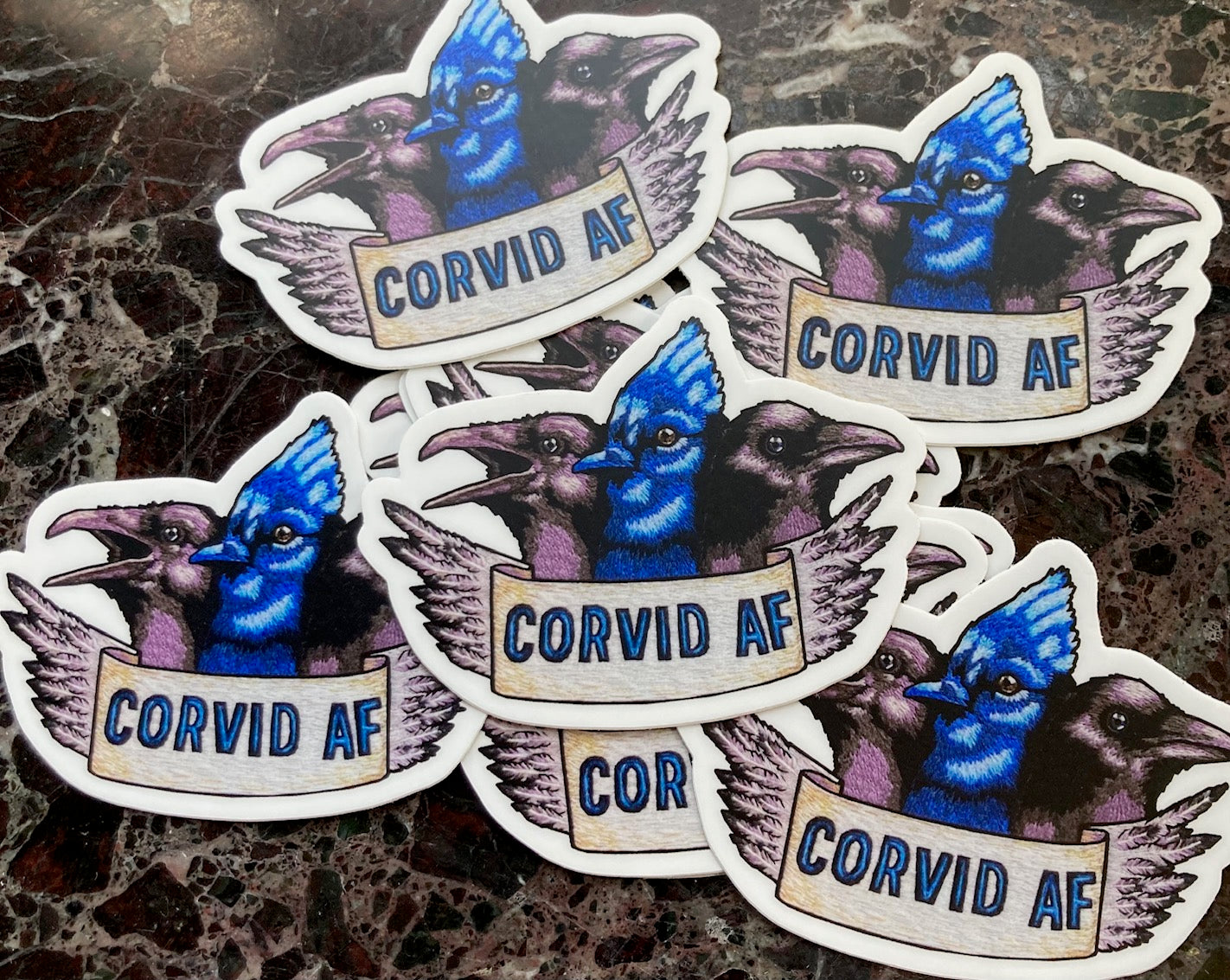 A pile of stickers sits atop a dark marble surface. The stickers depict an embroidery of the heads of three Corvids: a raven, a blue jay, and a crow. Below them is a banner that reads "CORVID AF" and the edge of the banner is feathers.