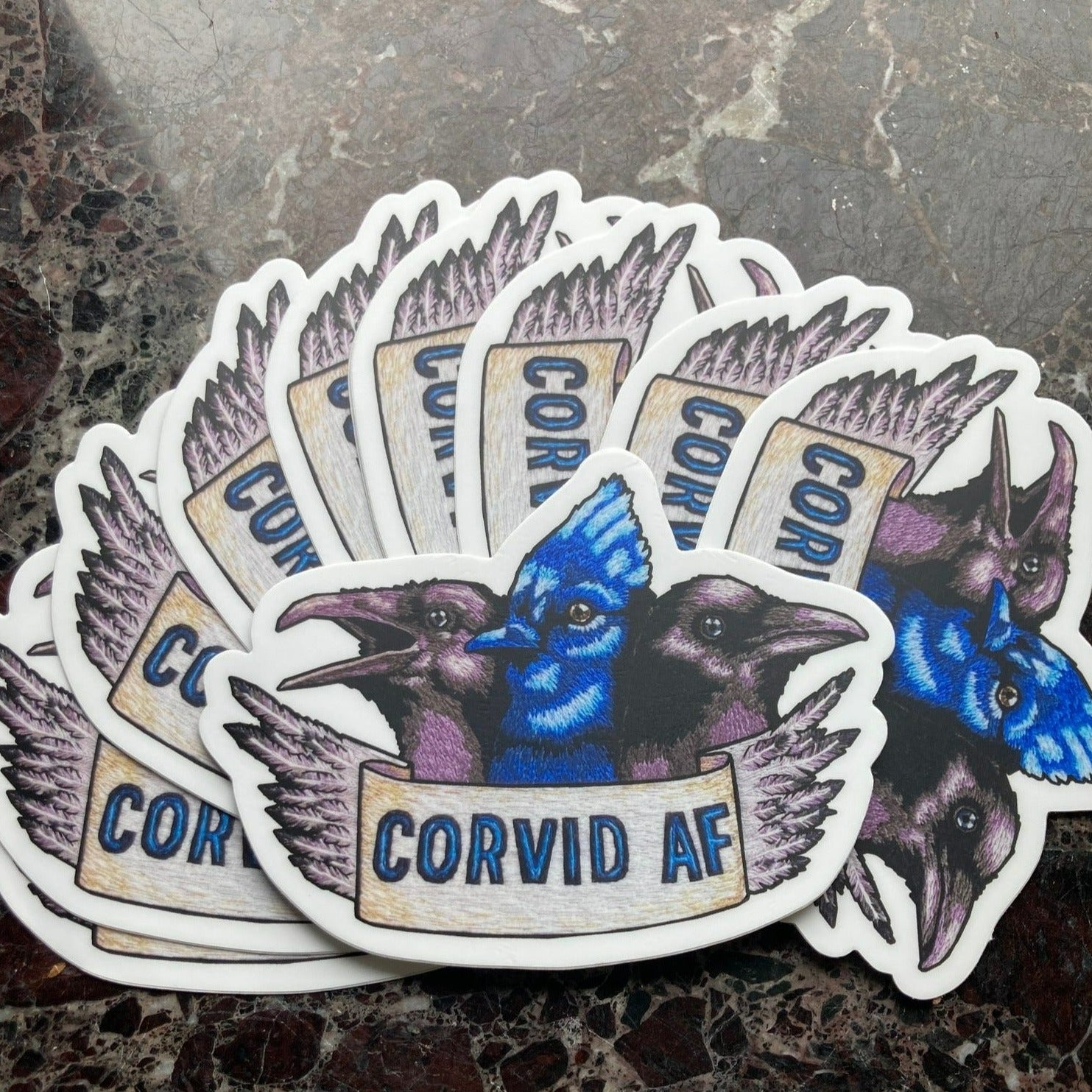 A pile of stickers sits atop a dark marble surface. The stickers depict an embroidery of the heads of three Corvids: a raven, a blue jay, and a crow. Below them is a banner that reads "CORVID AF" and the edge of the banner is feathers.