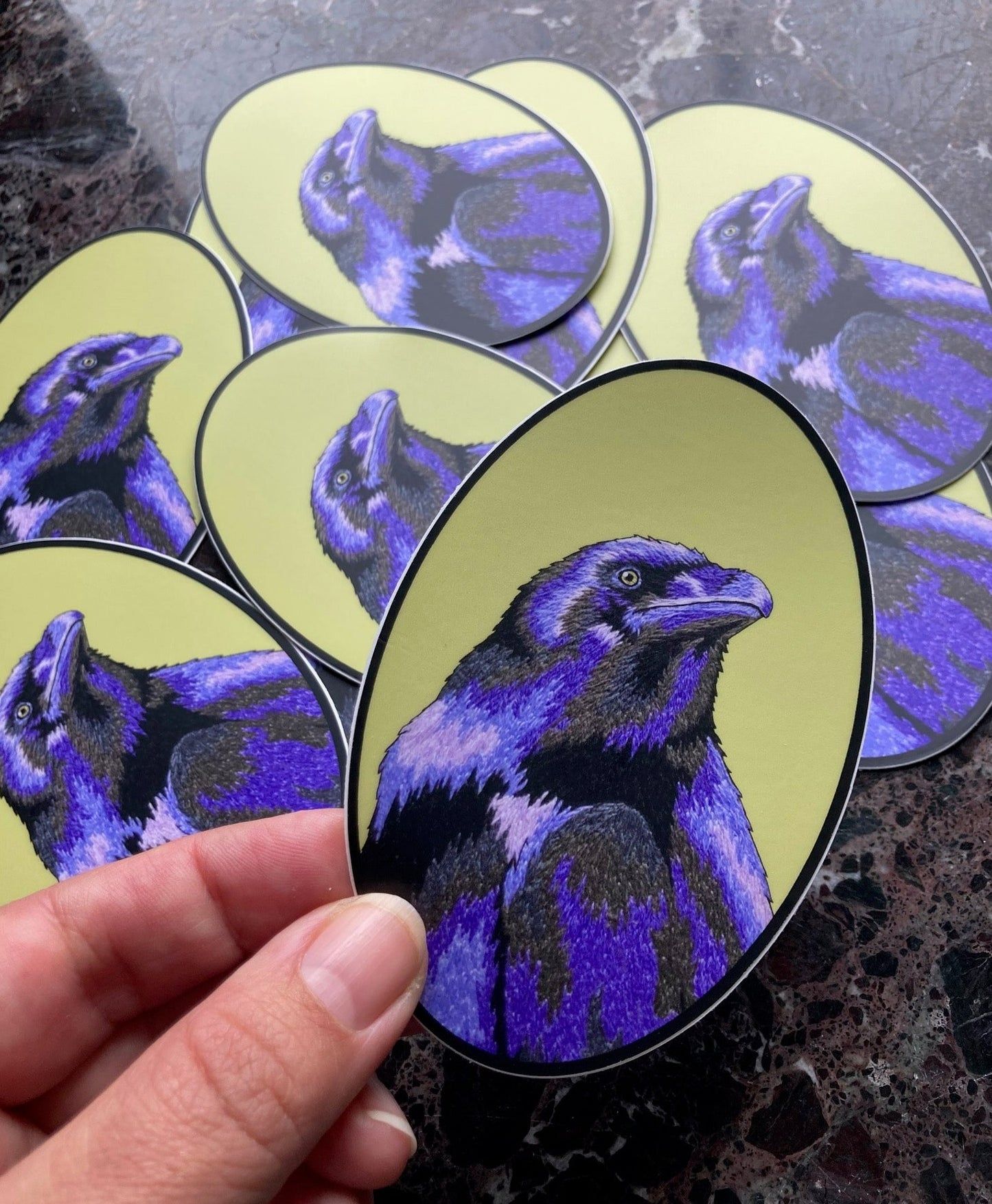 A pile of oval stickers sits atop a dark marble surface. The stickers depict an embroidered raven in deep periwinkle purples and greys against a green background. A hand holds one of the stickers up to the camera.