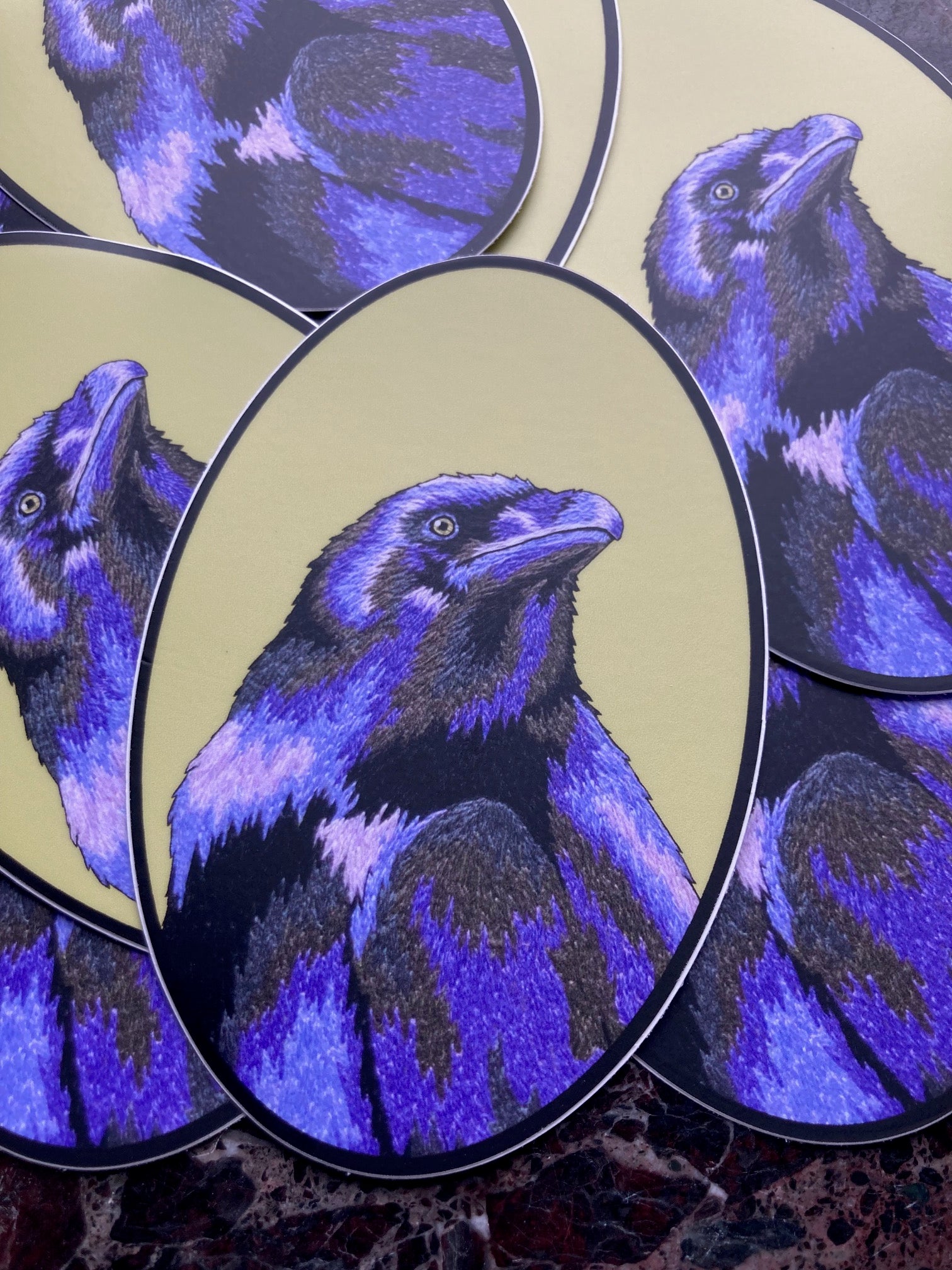 A pile of oval stickers sits atop a dark marble surface. The stickers depict an embroidered raven in deep periwinkle purples and greys against a green background.