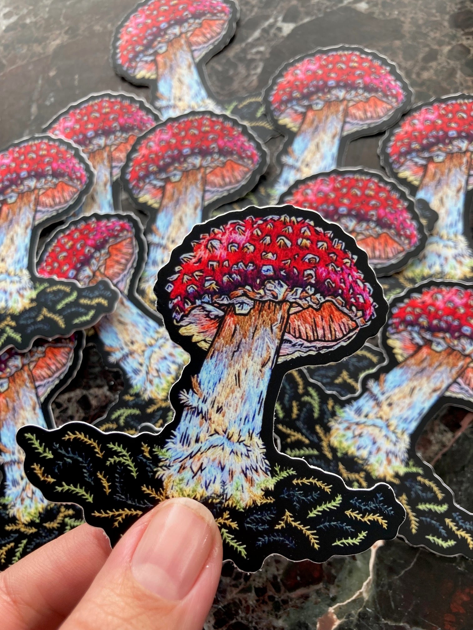 A hand holds up a sticker depicting a brightly colored amanita mushroom. Behind it is a scatter of other stickers on a flat surface.