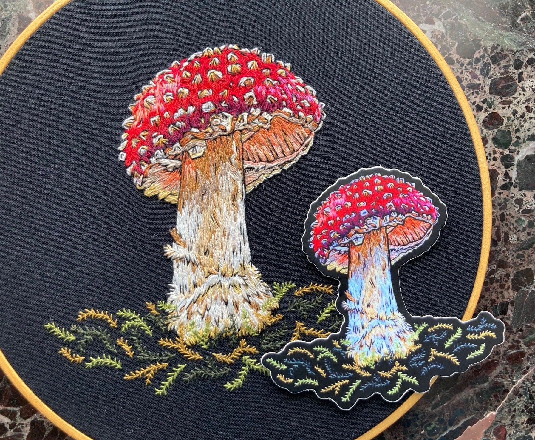 A sticker depicting a brightly colored embroidered amanita mushroom sits on the original embroidery of the same Amanita Mushroom.