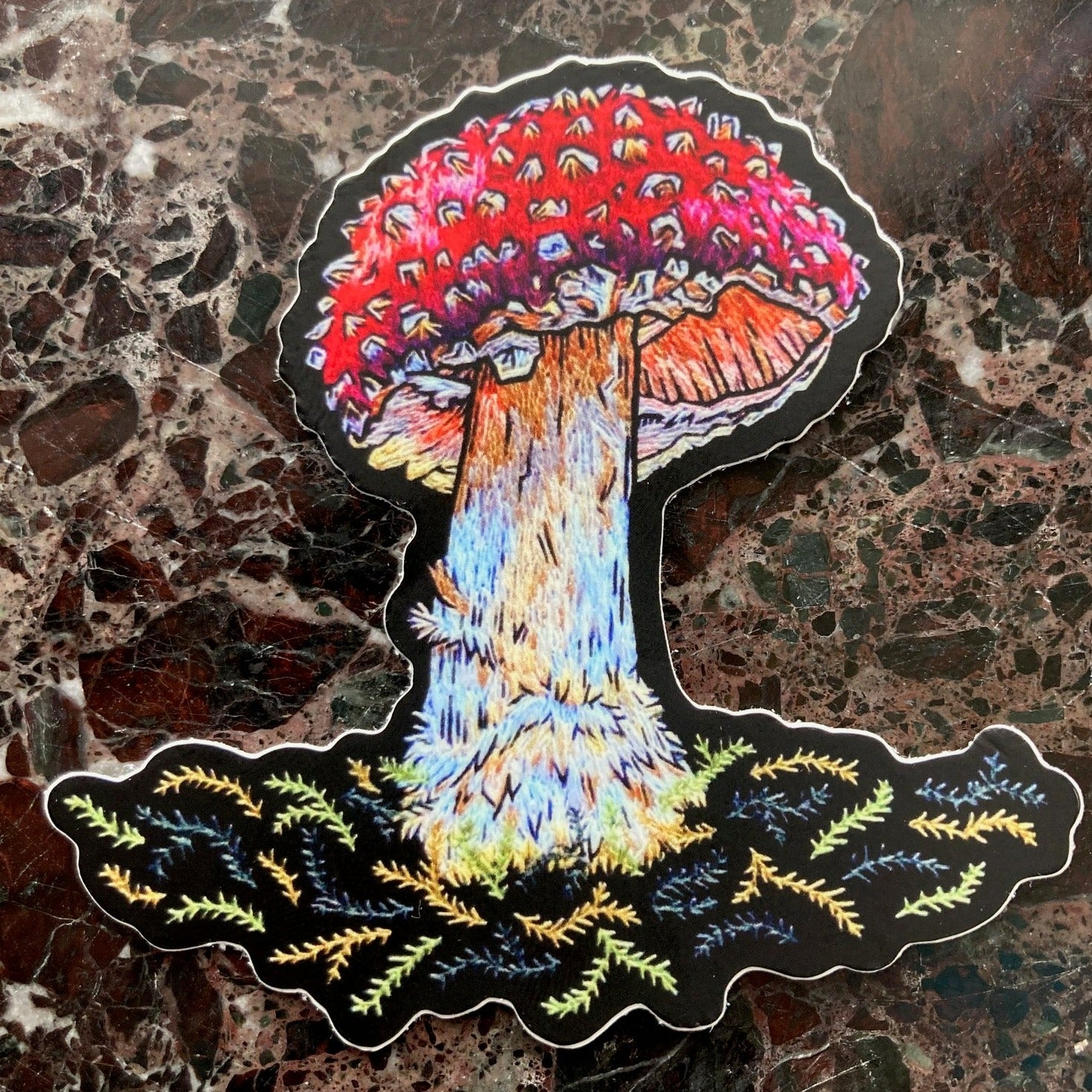 A sticker depicting a brightly colored embroidered amanita mushroom sits on a dark marble surface.