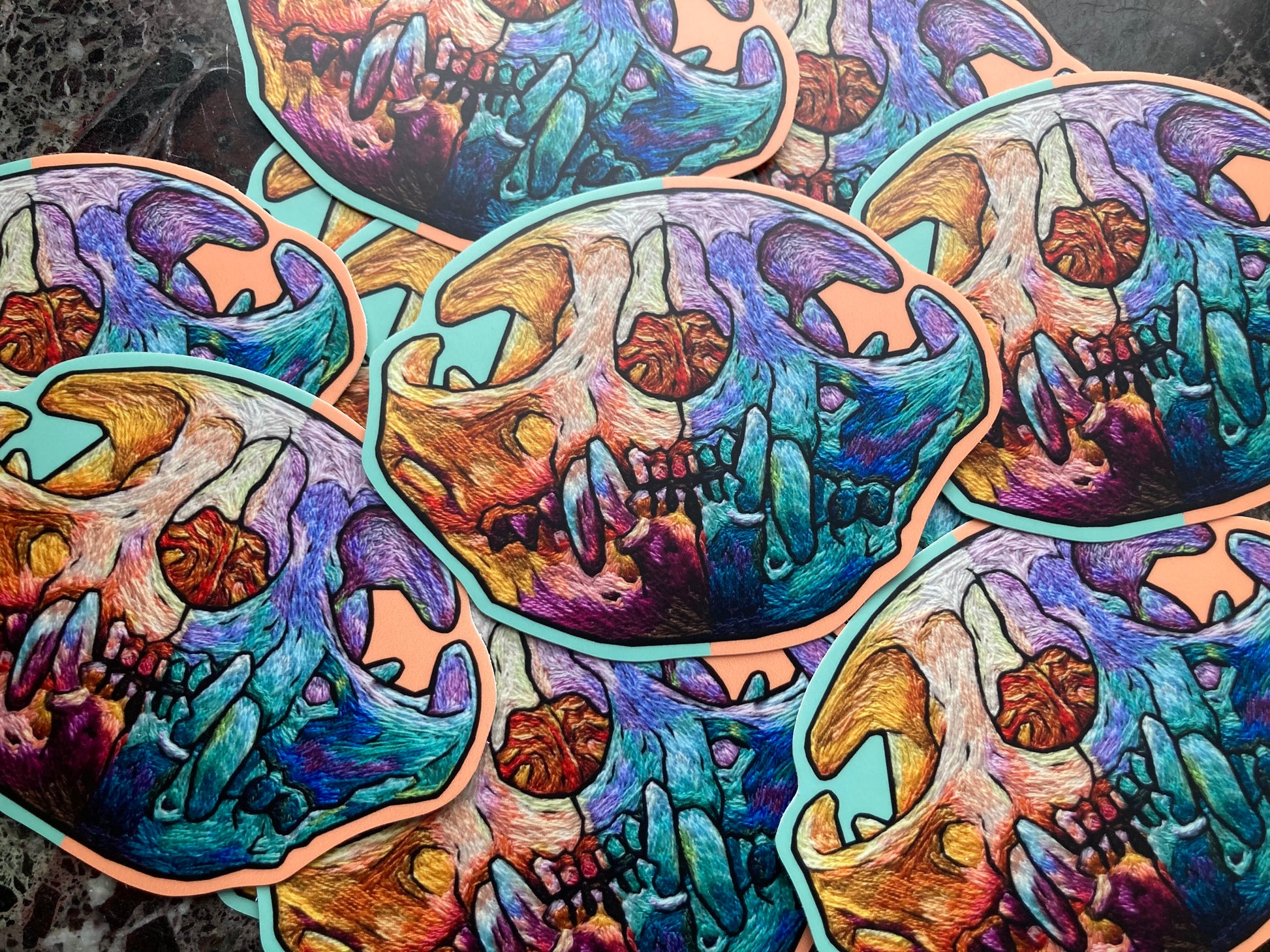 A pile of stickers sits on a dark surface. Each large sticker has a brightly colored embroidered cat skull printed on it.