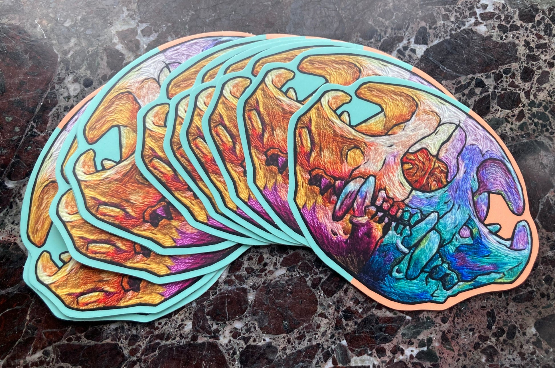 A pile of stickers sits on a dark surface. Each large sticker has a brightly colored embroidered cat skull printed on it.