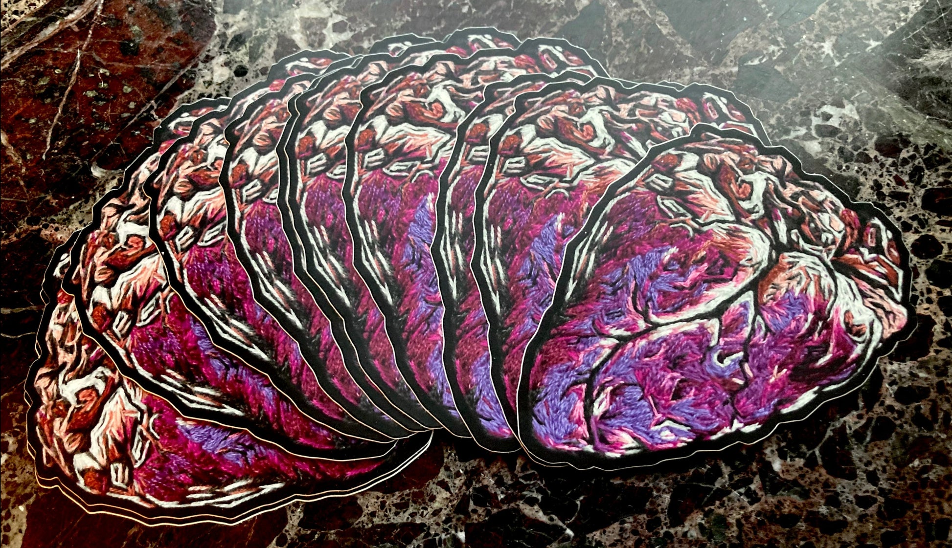A fanned out pile of stickers printed with a graphic embroidered anatomical heart are lying on a dark surface. The heart has bright pink and purple tones, with white and beige highlights, and a dark black outline.