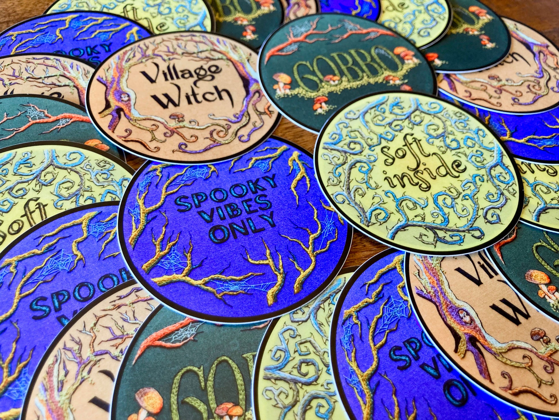 many round stickers based off of embroideries with various text like "Spooky vibes only" and "village witch" are arranged on a table, with four of them prominently laid out.