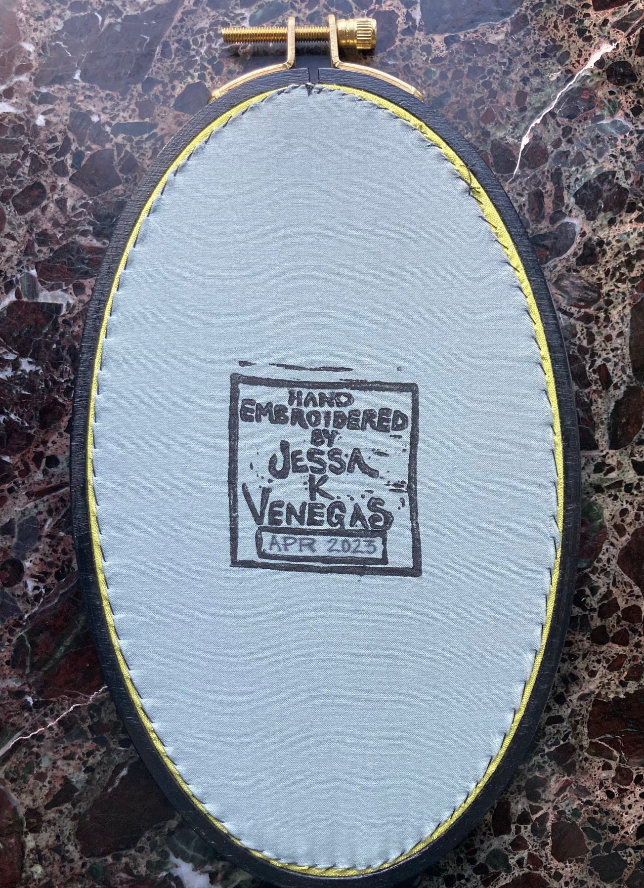 grey fabric is stretched into the back of an oval embroidery hoop. The fabric is printed with "Hand Embroidered by Jessa K Venegas" and the date April 2023 is written in.