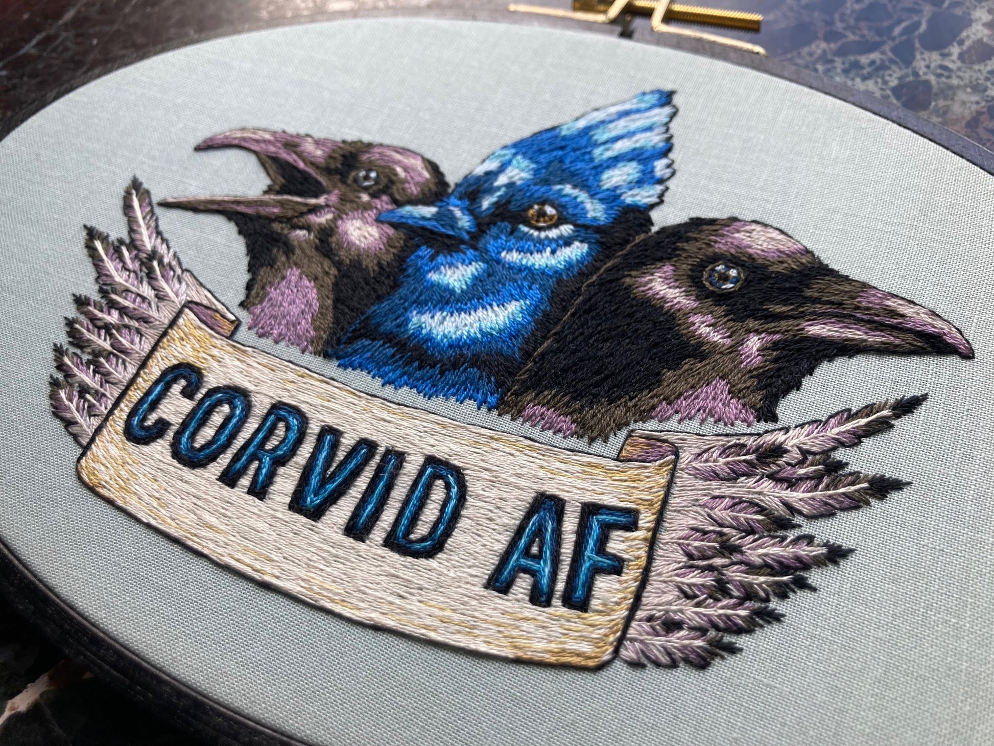 three birds are embroidered in an oval hoop. the outer birds are black and purple and the inner one is bright blue. a banner at the bottom says "Corvid AF"