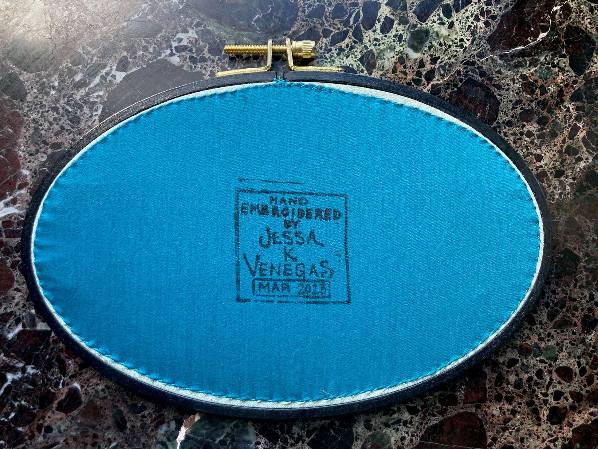 the back of an embroidery hoop is pictured, with teal fabric stitched onto it. There is a printed label on the fabric reading "Hand Embroidered by Jessa K Venegas" and below it is written "March 2023" in ink.