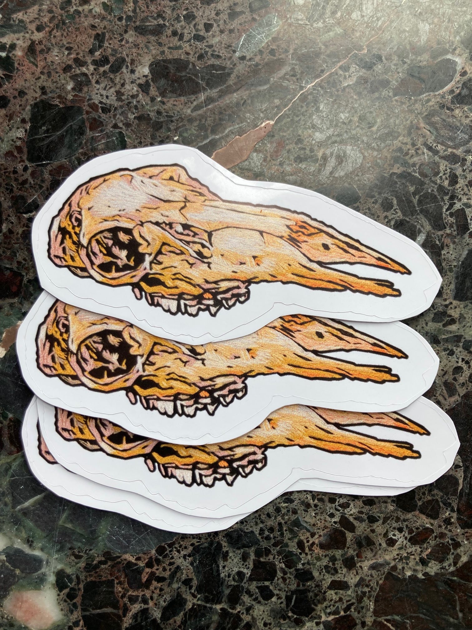 a pile of stickers with an image of an embroidered deer skull with no lower jaw sits on a dark surface.