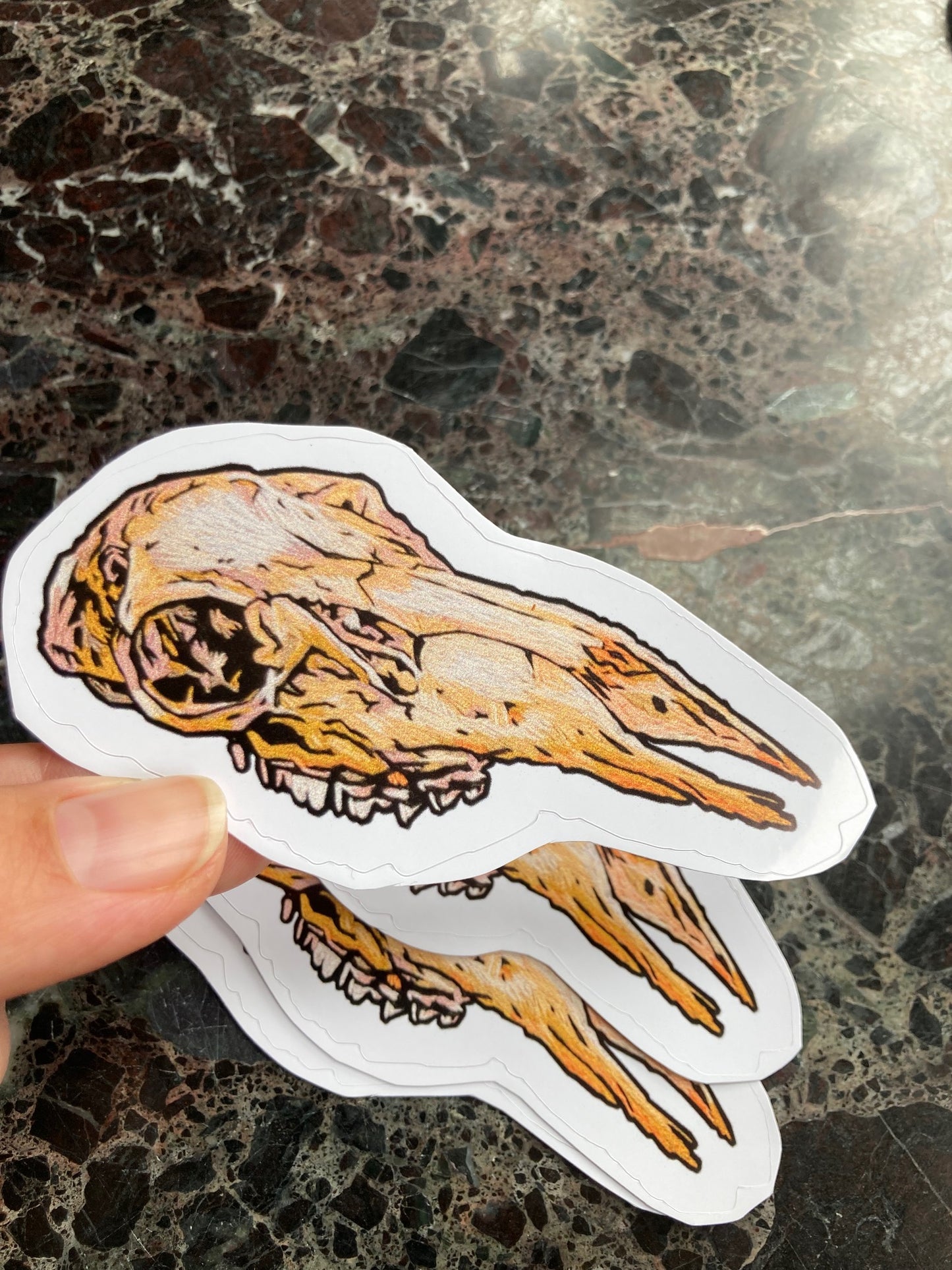 a pile of stickers with an image of an embroidered deer skull with no lower jaw sits on a dark surface. a hand is holding one up to the camera.