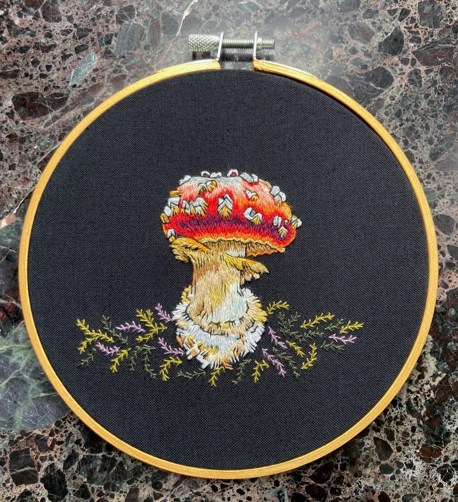 A brightly colored amanita mushroom with a red cap is embroidered on black fabric with green and lavender moss. The fabric is stretched into a gold round embroidery hoop.