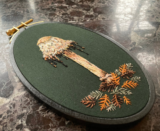 An embroidery hoop sits on a black marble background. It features a hand embroidered shaggy mane mushroom with a drippy cap and some stitched redwood leaves, on a dark green fabric.