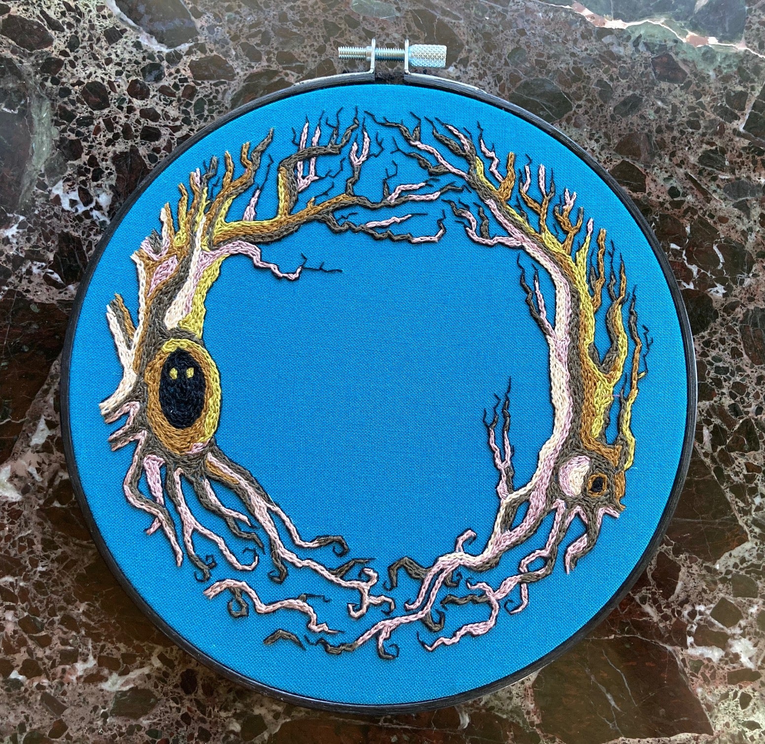 An embroidery hoop sits on a black marble background. It features two gnarled trees stitched in brown and lilac and green. One tree has little eyes peering out from a dark knot.