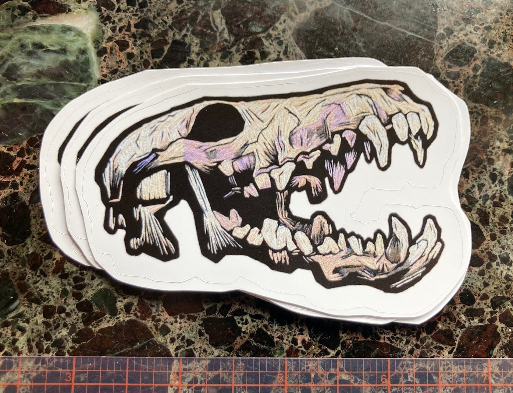 a sticker depicting an embroidered coyote skeleton with an open mouth and lots of teeth sits on a dark surface. The Embroidered skull is beige and lavender and black tones with lots of shadows. A visible ruler shows the sticker to be a little less than 4 inches long.