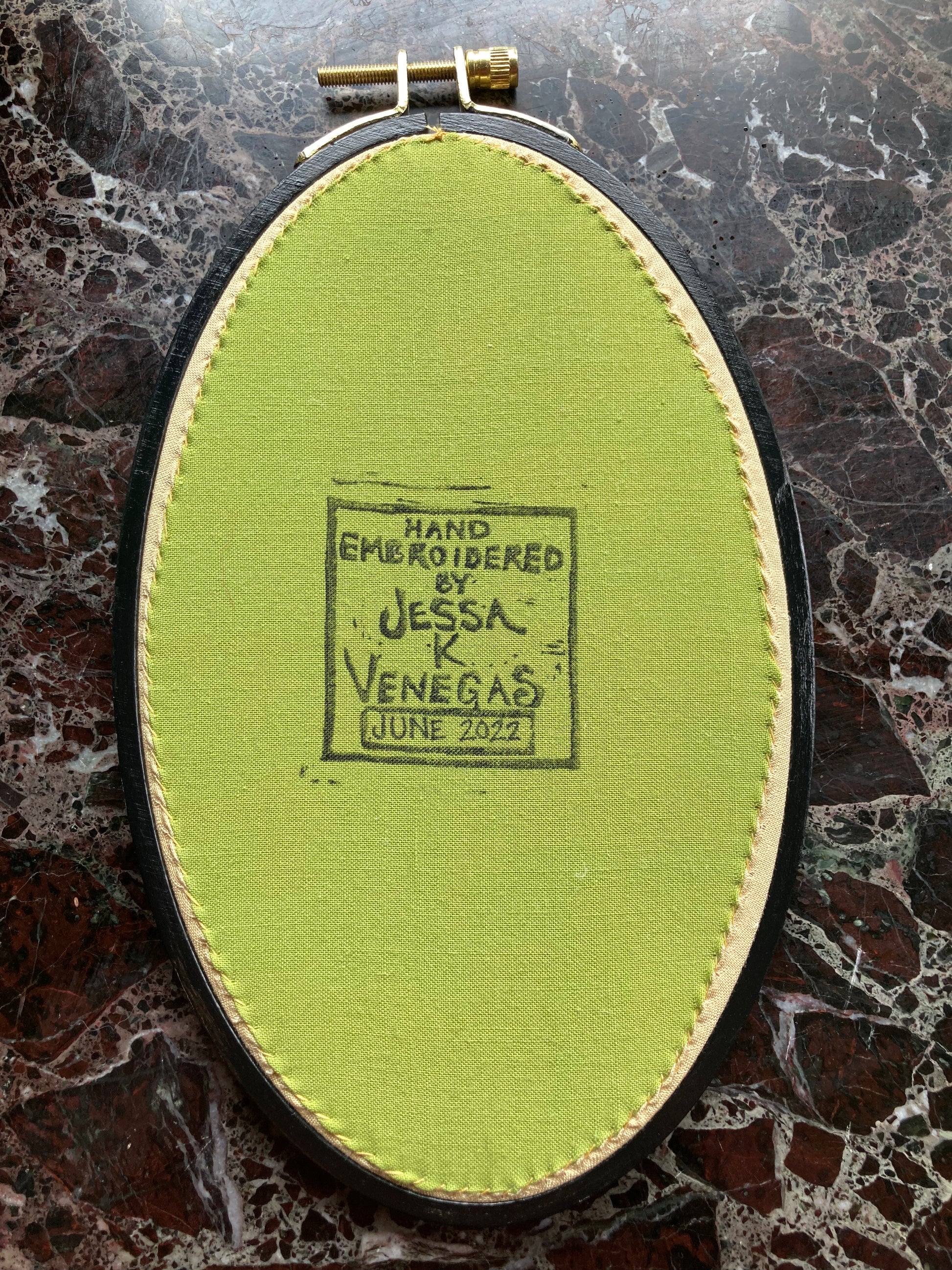 Lime green fabric is stitched taut into the back of a black oval embroidery hoop. The fabric is stamped with a label reading "Hand embroidered by Jessa K Venegas". Below the label "June 2022" is written in ink. 