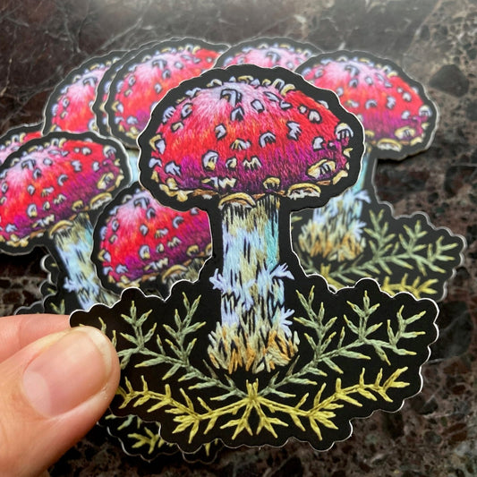 a pile of stickers sits on a dark surface. The stickers depict an embroidery of a mushroom with a red cap and white spots above some decorative stitched green moss. A hand holds one up to the camera. 