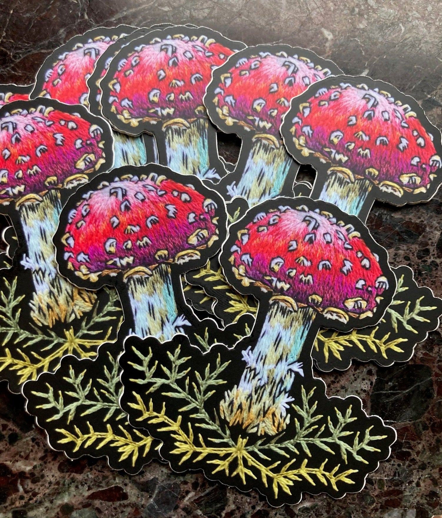 a pile of stickers sits on a dark surface. The stickers depict an embroidery of a mushroom with a red cap and white spots above some decorative stitched green moss. 