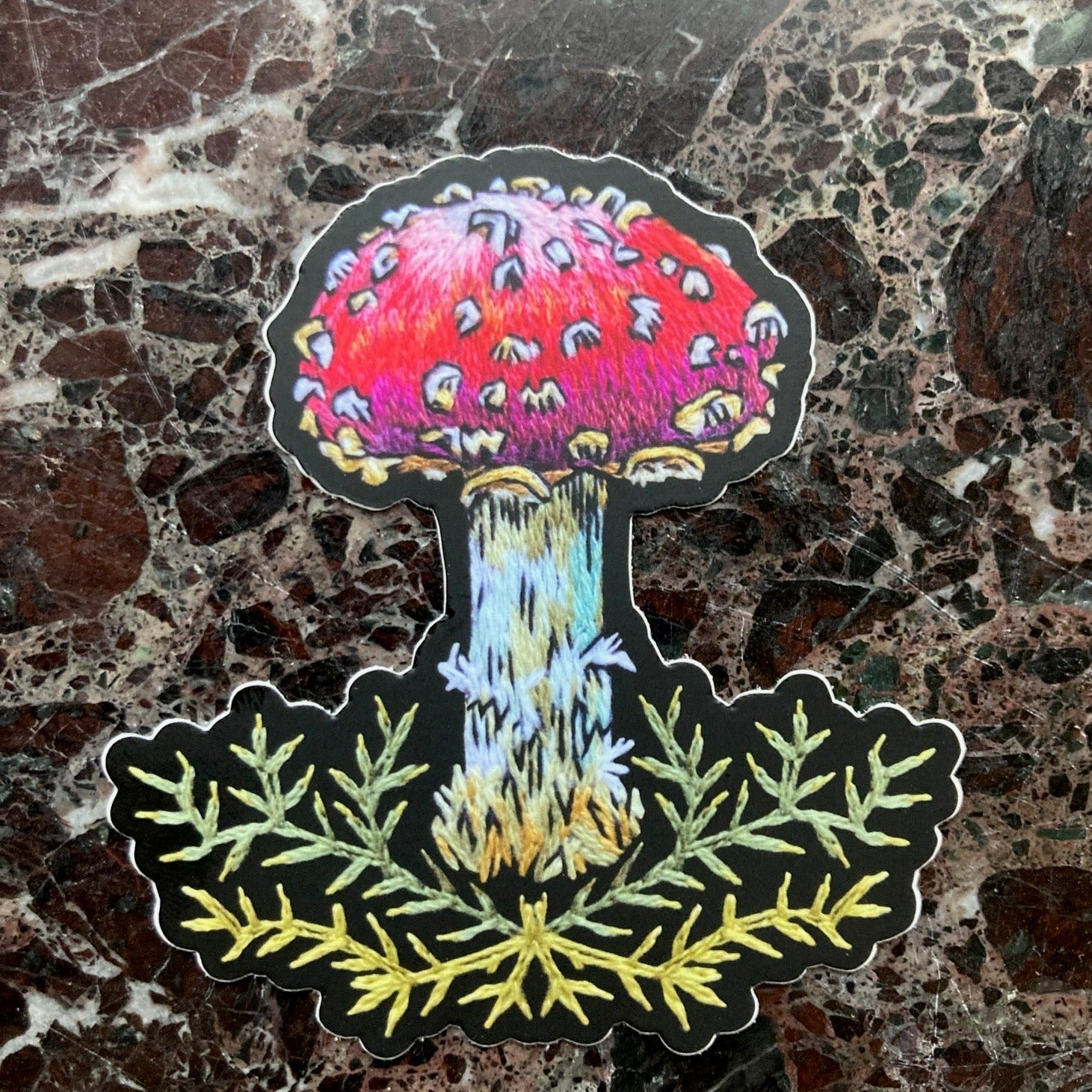 a single sticker depicting an embroidery of a mushroom with a red cap and white spots above some decorative stitched green moss sits on a dark marble surface.
