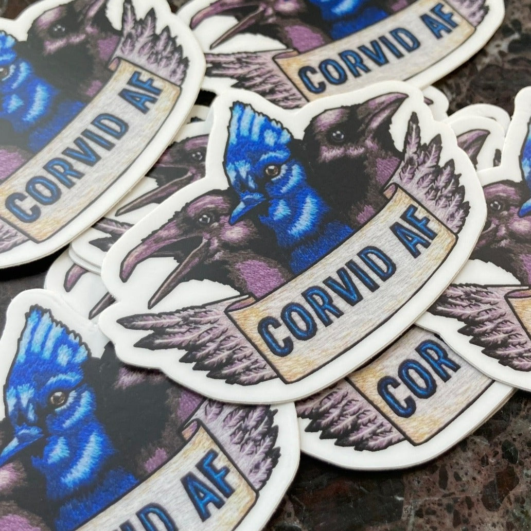 A pile of stickers sits atop a dark marble surface. The stickers depict an embroidery of the heads of three Corvids: a raven, a blue jay, and a crow. Below them is a banner that reads "CORVID AF" and the edge of the banner is feathers.