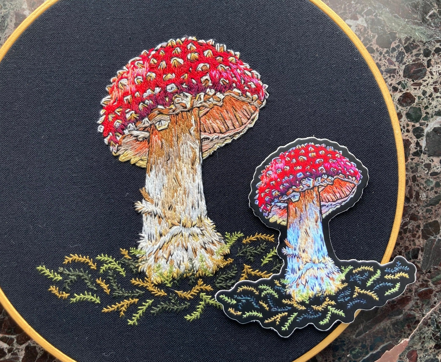 A sticker depicting a brightly colored embroidered amanita mushroom sits on the original embroidery of the same Amanita Mushroom. 