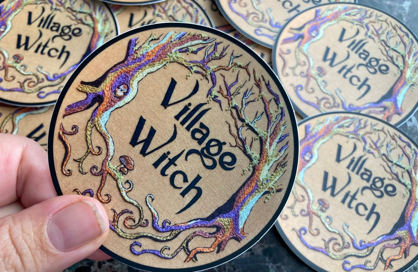 a pile of stickers lays on a dark surface. The stickers depict an embroidery of colorful trees with moss on a tan background and the words "Village Witch" in the center. There are mushrooms and a tiny owl in the trees.