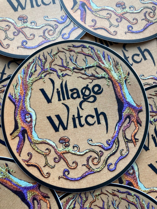 a pile of stickers lays on a dark surface. The stickers depict an embroidery of colorful trees with moss on a tan background and the words "Village Witch" in the center. There are mushrooms and a tiny owl in the trees.