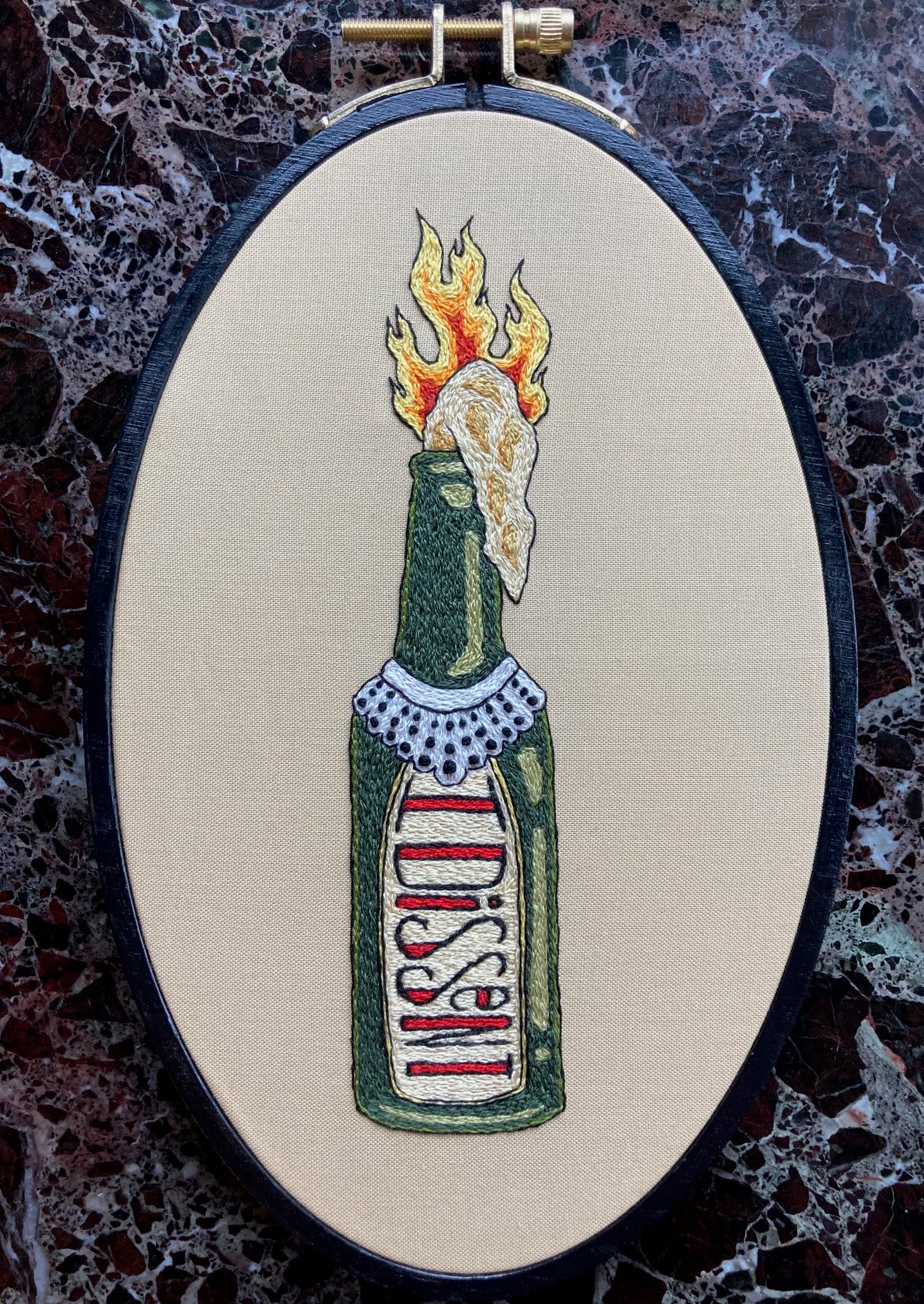 A flaming molotov cocktail bottle with a lace collar around the neck in the style of Ruth Bader Ginsburg is embroidered onto tan fabric in a black oval hoop. The label of the bottle reads "I DISSENT" in decorative font. The hoop sits upon a dark marble surface.