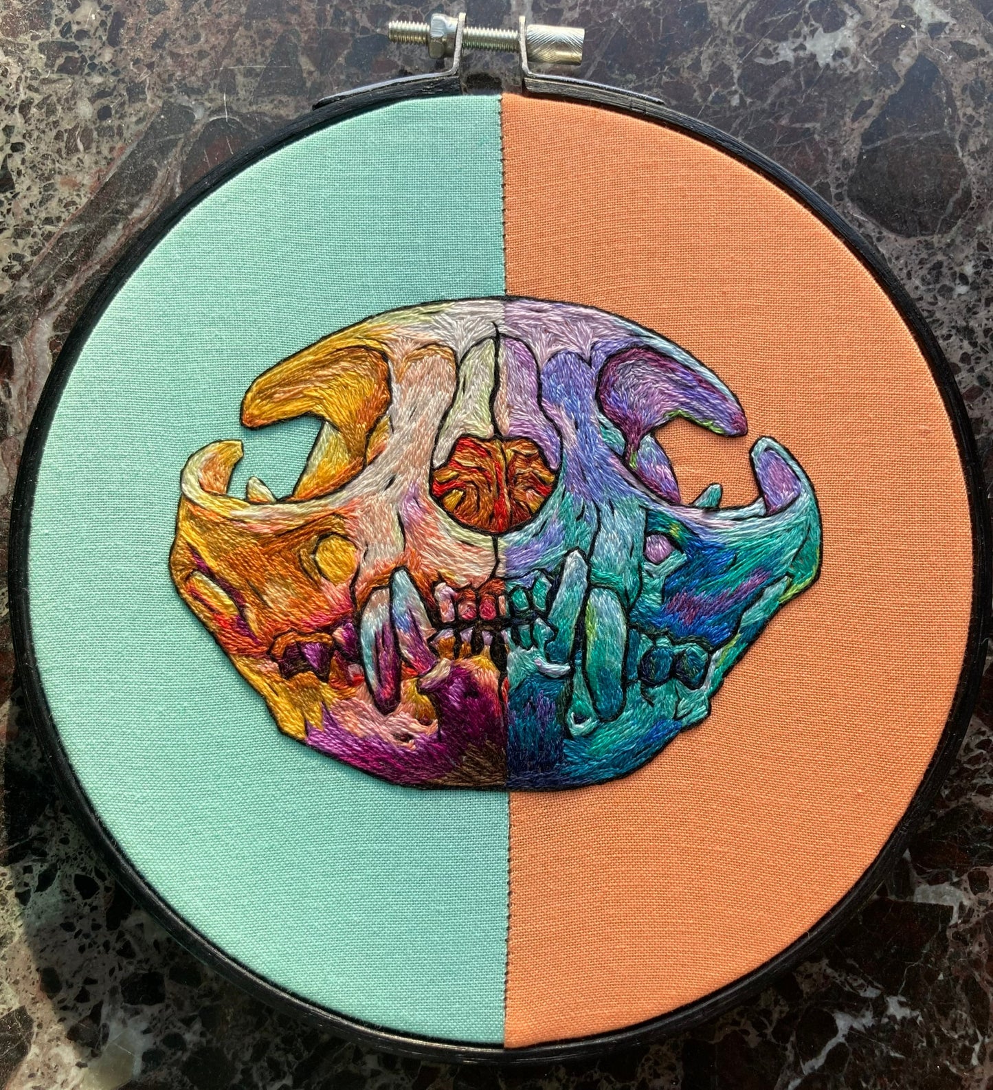 a vibrantly colored embroidered cat skull in a black hoop sits on a black marble surface. The fabric in the hoop is aqua on the left side and apricot on the right, and the stitching mirrors that split with warm tones on the left and cool tones on the right. 