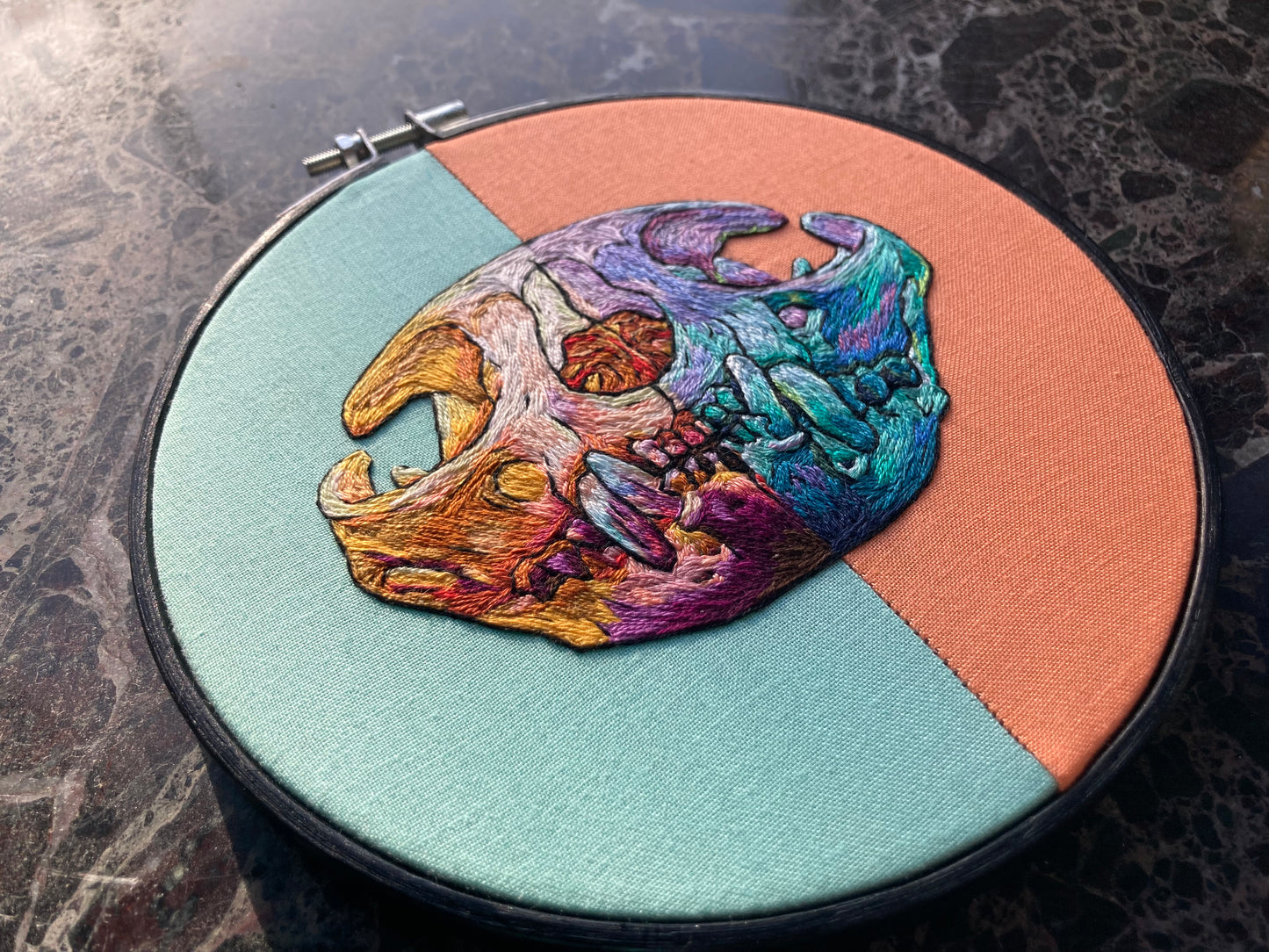 a vibrantly colored embroidered cat skull in a black hoop sits on a black marble surface. The fabric in the hoop is aqua on the left side and apricot on the right, and the stitching mirrors that split with warm tones on the left and cool tones on the right. 