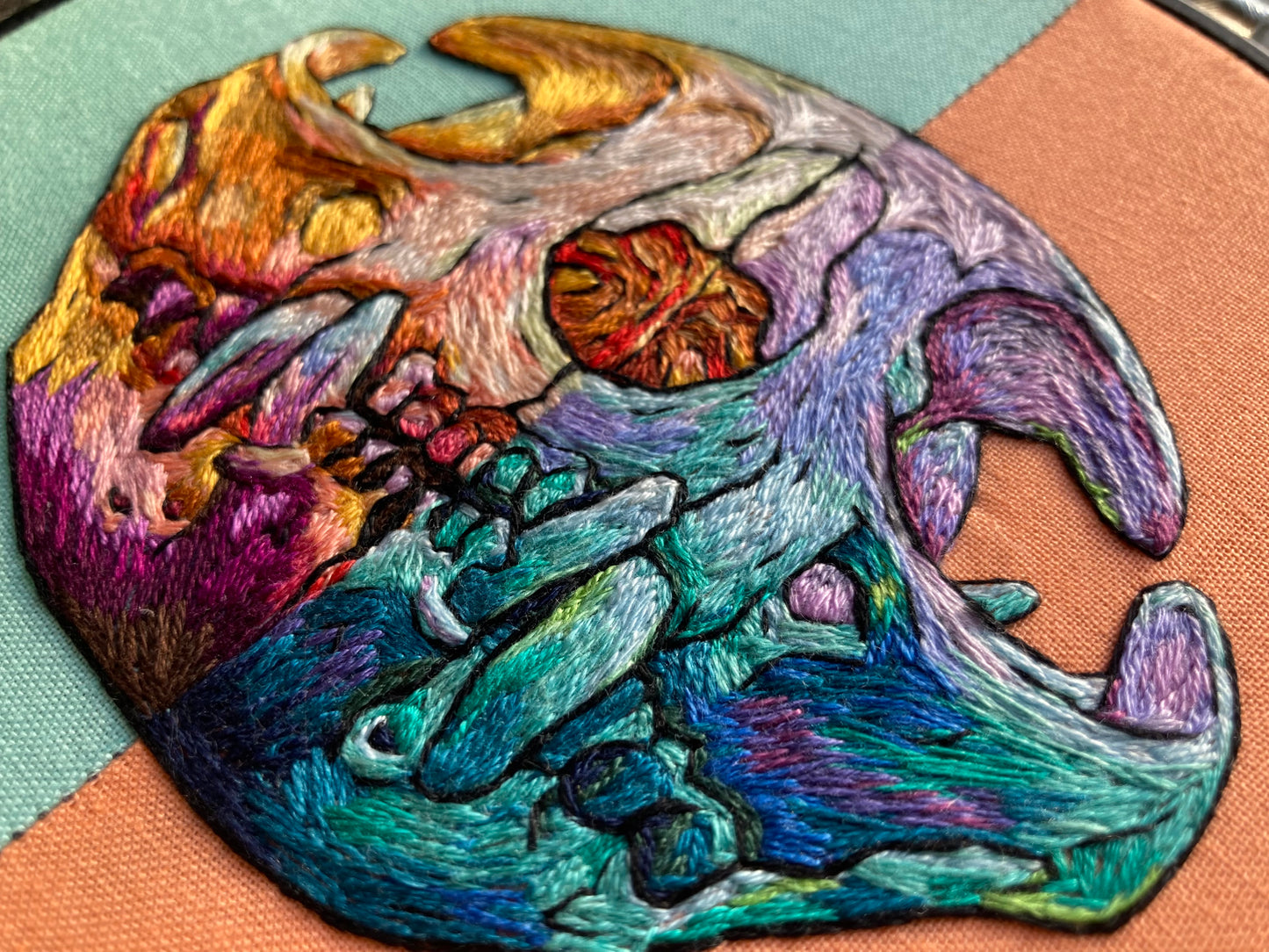 a vibrantly colored embroidered cat skull in a black hoop. The photo is taken very close up and you can see the individual threads. The fabric in the hoop is aqua on the left side and apricot on the right, and the stitching mirrors that split with warm tones on the left and cool tones on the right. 