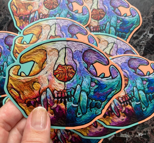 A hand holds up a large sticker with a brightly colored embroidered cat skull printed on it. There are a pile of the stickers on the dark surface below.