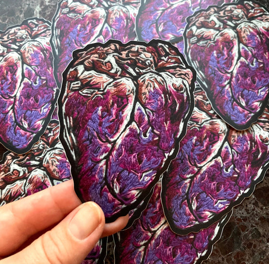 A pile of stickers printed with a graphic embroidered anatomical heart are lying on a dark surface. a hand holds up one sticker to the camera.