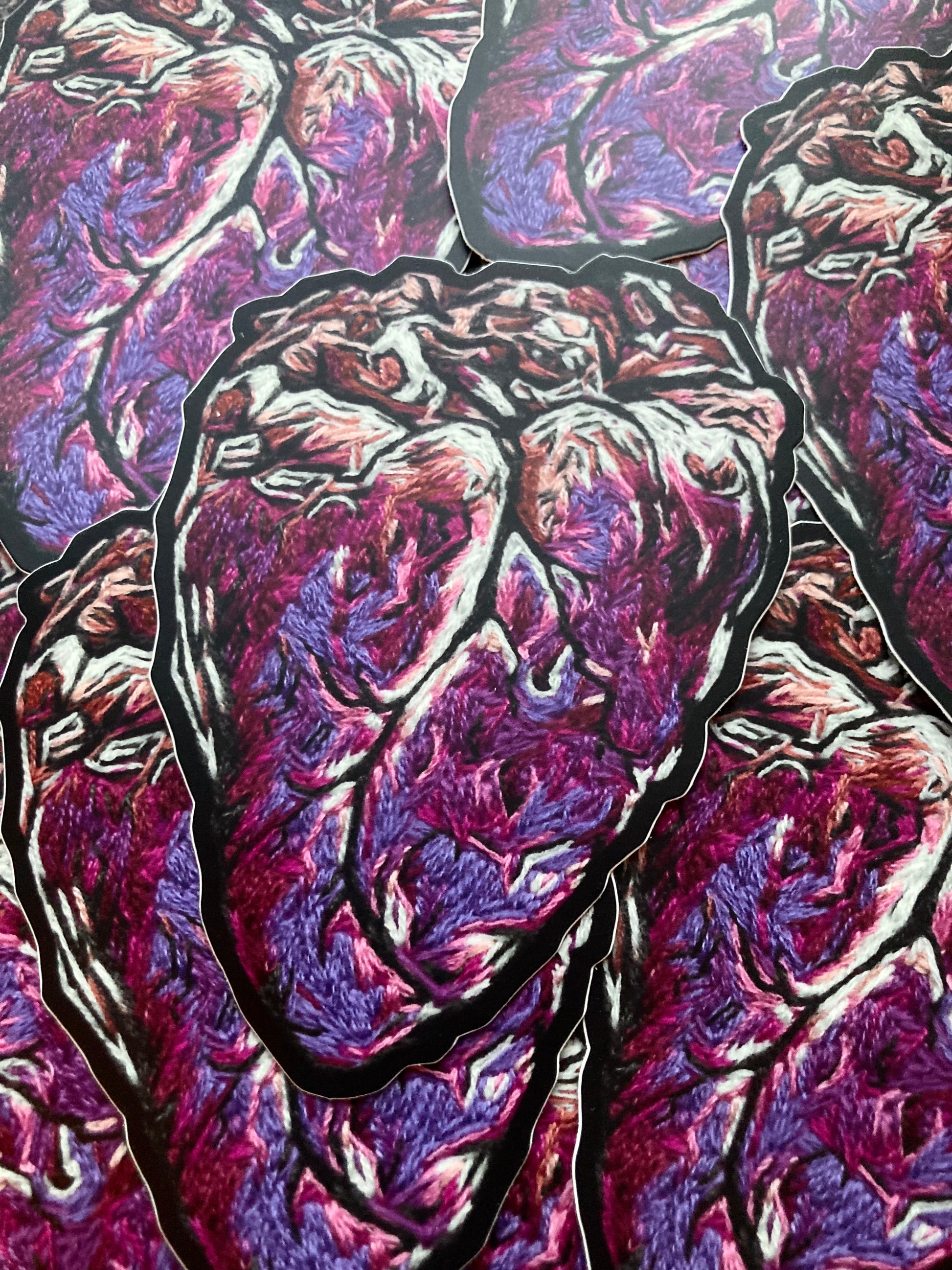 A pile of stickers printed with a graphic embroidered anatomical heart are lying on a dark surface. The heart has bright pink and purple tones, with white and beige highlights, and a dark black outline. 