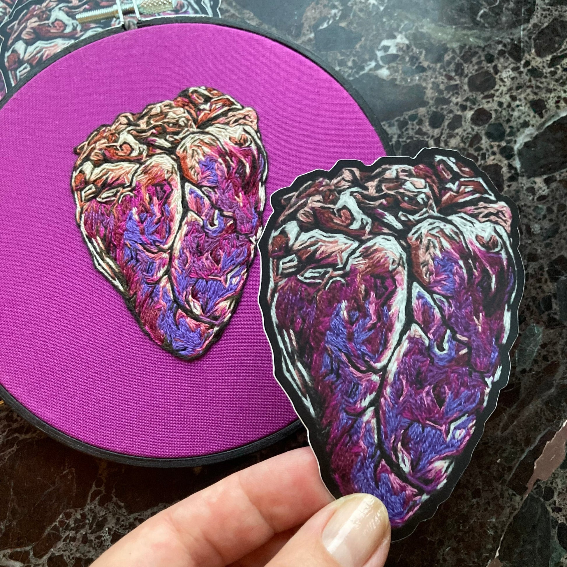 a hand holds up a printed sticker to the camera. Below it is the original embroidery of an anatomical heart on a bright fuchsia fabric. The sticker is a copy of this same design. 