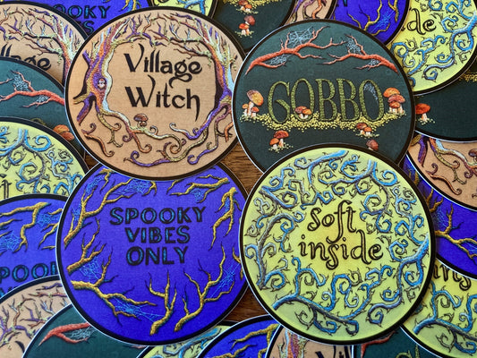 many round stickers based off of embroideries with various text like "Spooky vibes only" and "village witch" are arranged on a table, with four of them prominently laid out.