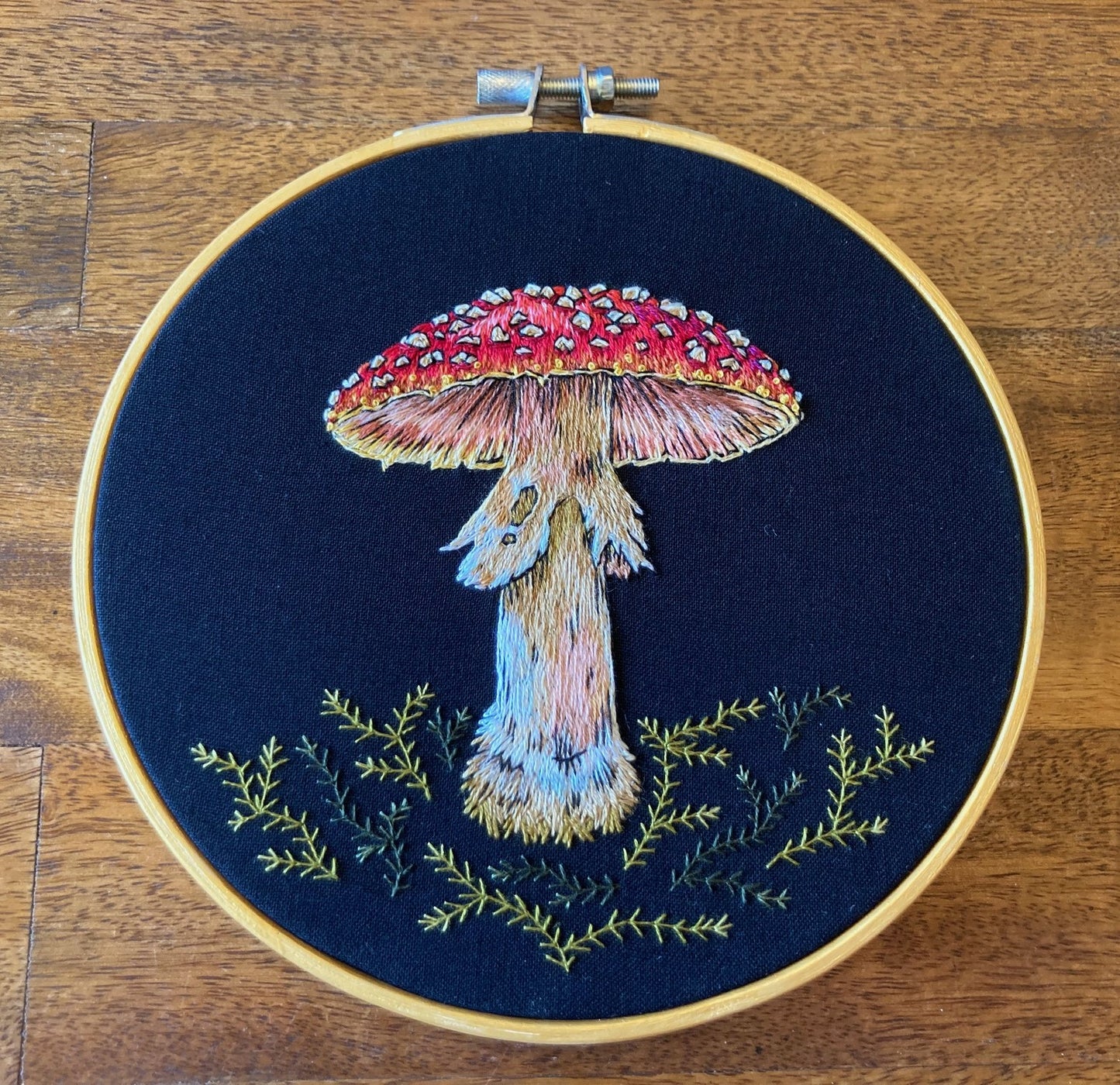an embroidered mushroom with a spotted red cap and a shaggy stem stands among green embroidered moss on a black fabric in a round gold hoop