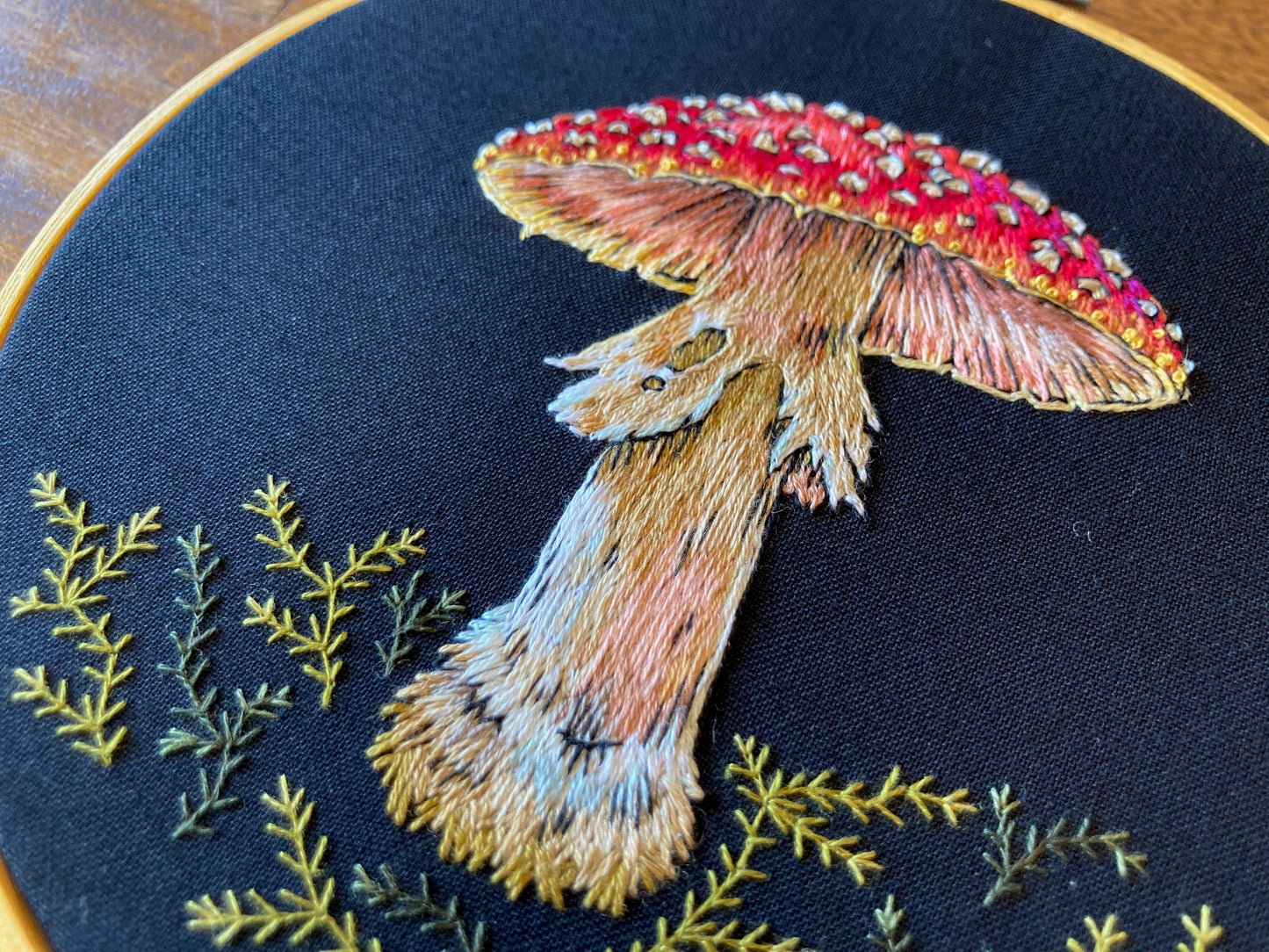 an embroidered mushroom with a spotted red cap and a shaggy stem stands among green embroidered moss on a black fabric in a round gold hoop