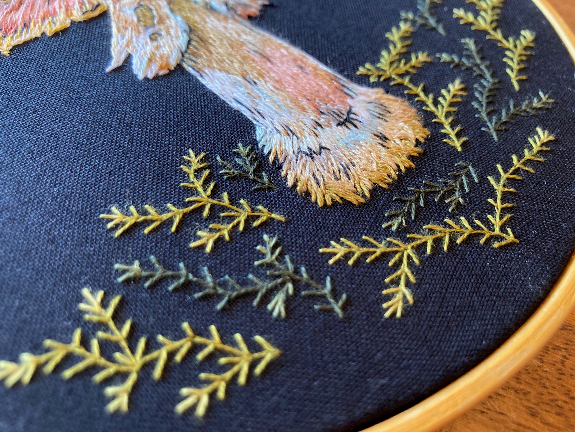 A closeup of an embroidered mushroom in which you can see only the stem among green embroidered moss on a black fabric in a round gold hoop