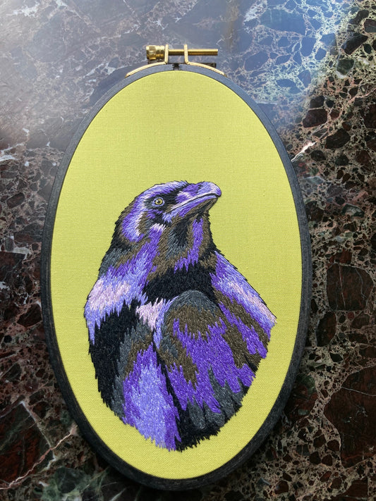 An embroidered raven stitched in black and purple threads looks up and to the right against a bright green background in a black painted oval hoop