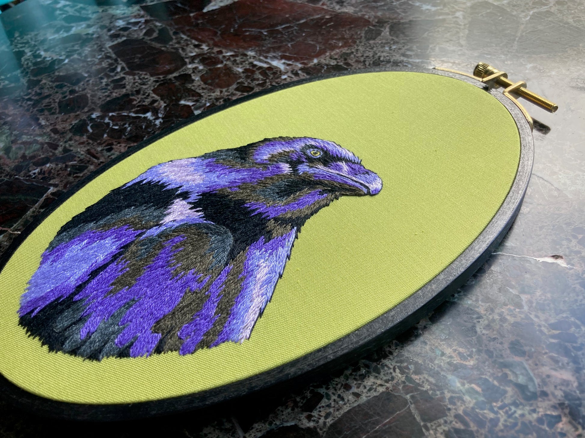 An embroidered raven stitched in black and purple threads looks up and to the right against a bright green background in a black painted oval hoop. The picture is taken at an extreme angle to capture the light in the threads