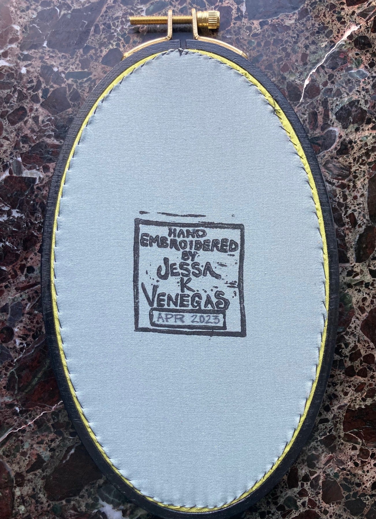 grey fabric is stretched into the back of an oval embroidery hoop. The fabric is printed with "Hand Embroidered by Jessa K Venegas" and the date April 2023 is written in. 