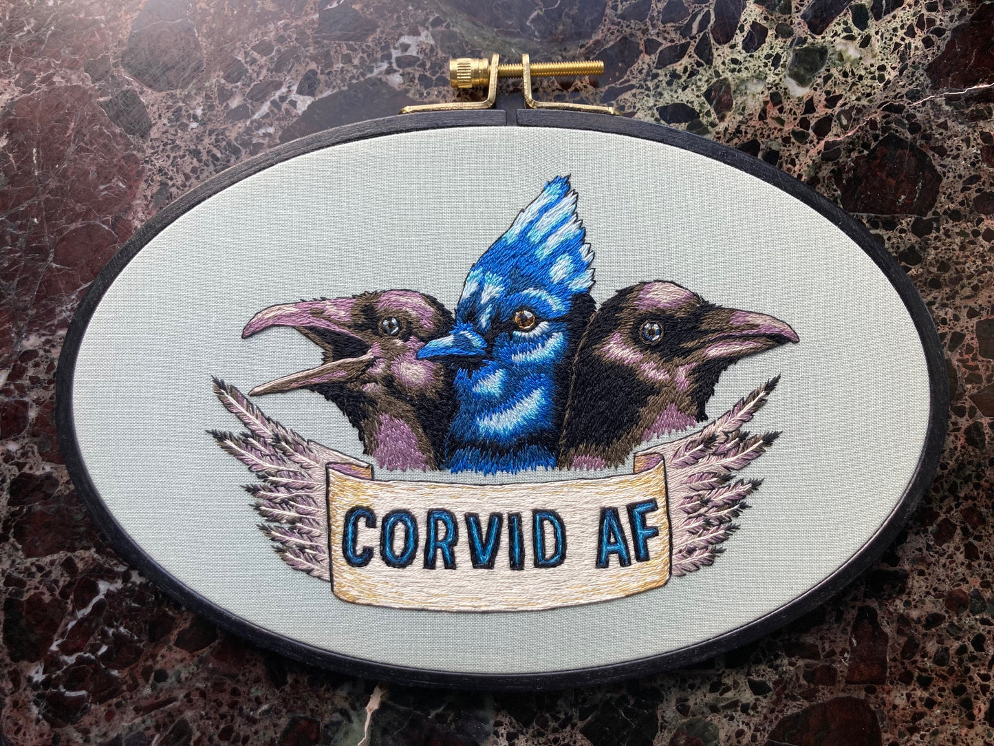 three birds are embroidered in an oval hoop. the outer birds are black and purple and the inner one is bright blue. a banner at the bottom says "Corvid AF"