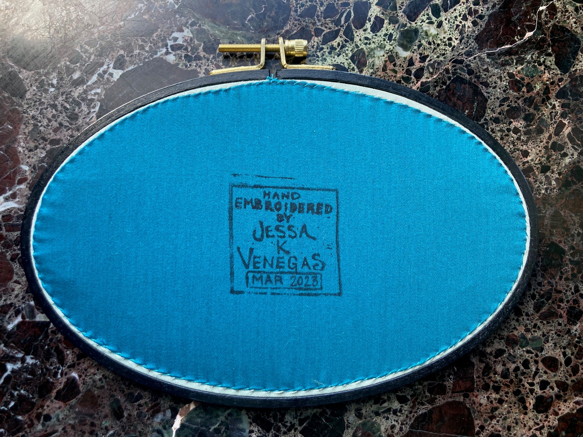 the back of an embroidery hoop is pictured, with teal fabric stitched onto it. There is a printed label on the fabric reading "Hand Embroidered by Jessa K Venegas" and below it is written "March 2023" in ink. 