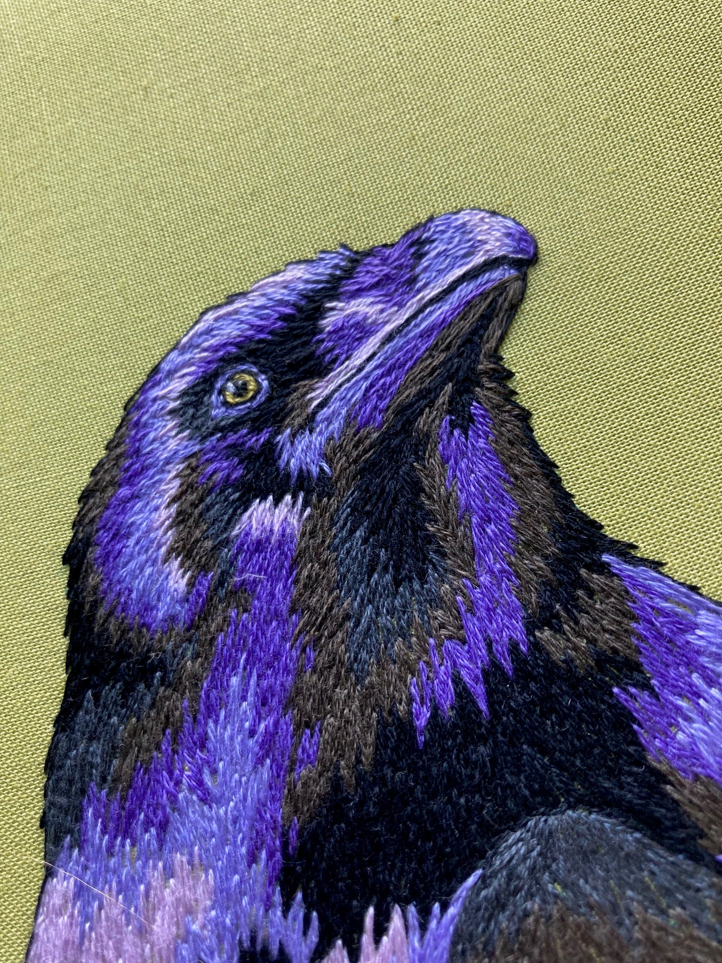 An embroidered raven stitched in black and purple threads looks up and to the right against a bright green background 