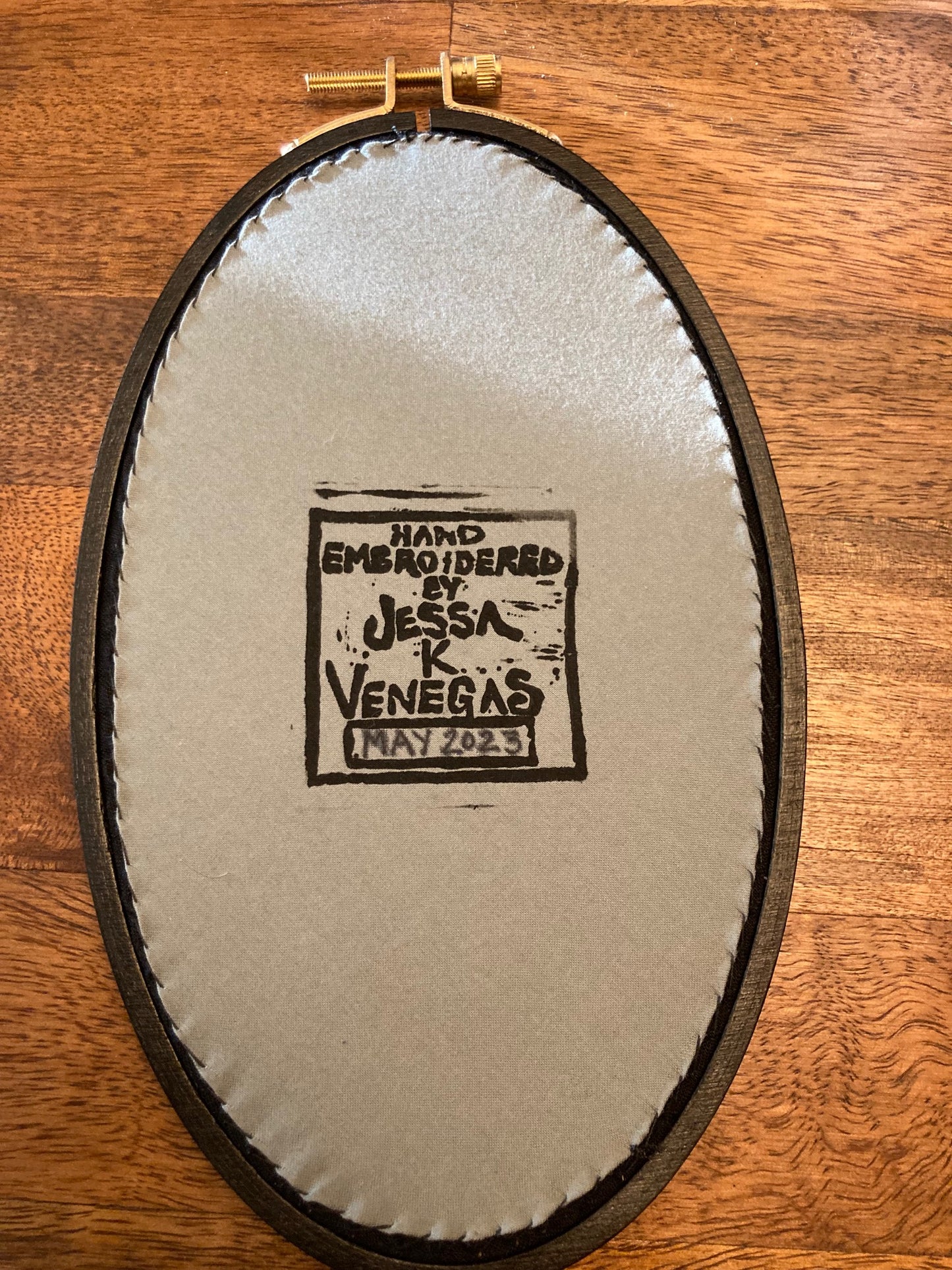 An oval embroidery hoop is backed with taut grey fabric that has a printed label reading "Hand Embroidered by Jessa K Venegas" and May 2023 is written below in ink. 