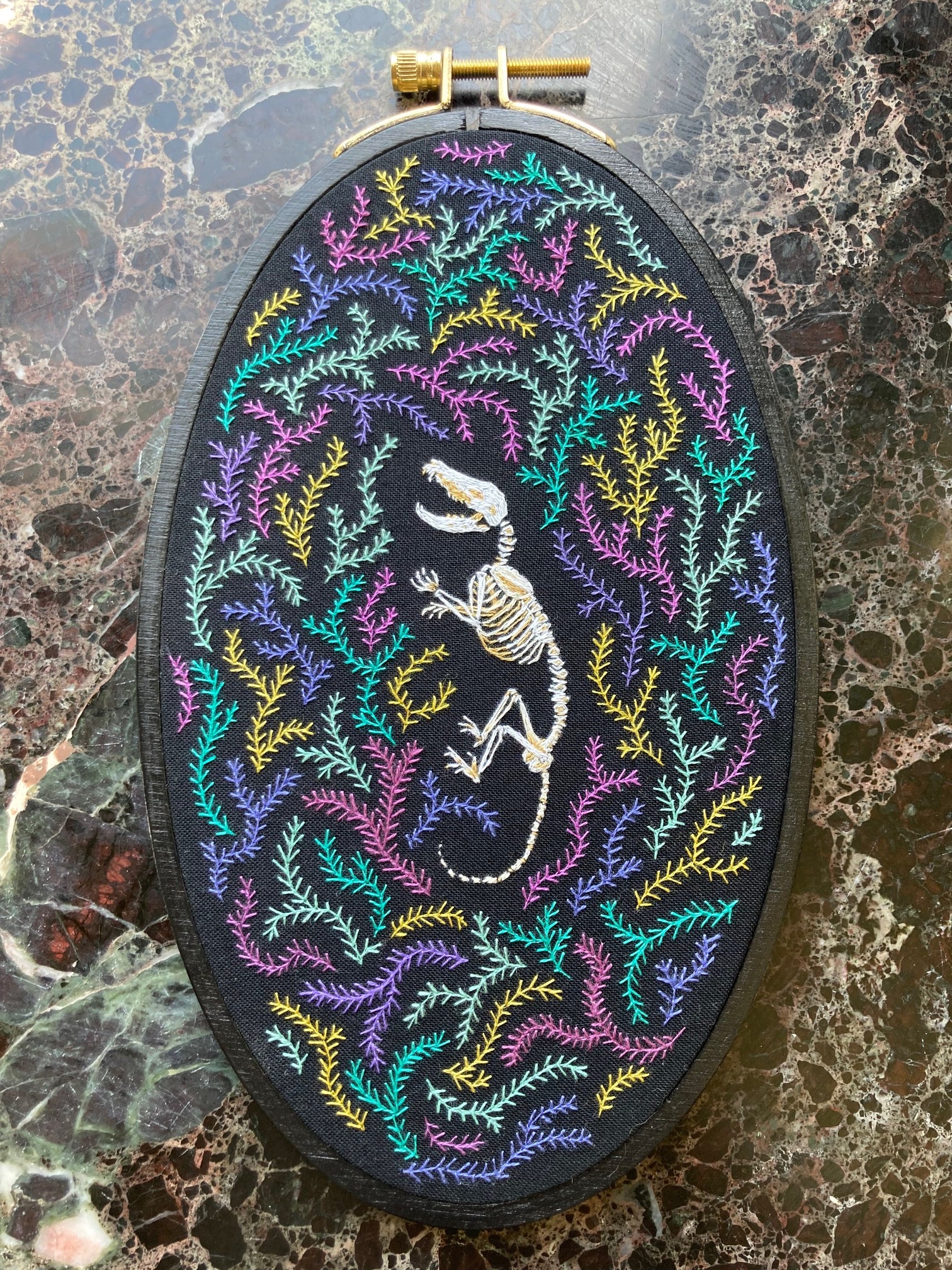 In a black oval hoop, an embroidered opossum skeleton rests among colorful pink and teal and golden stitched moss or fern against a black background. 